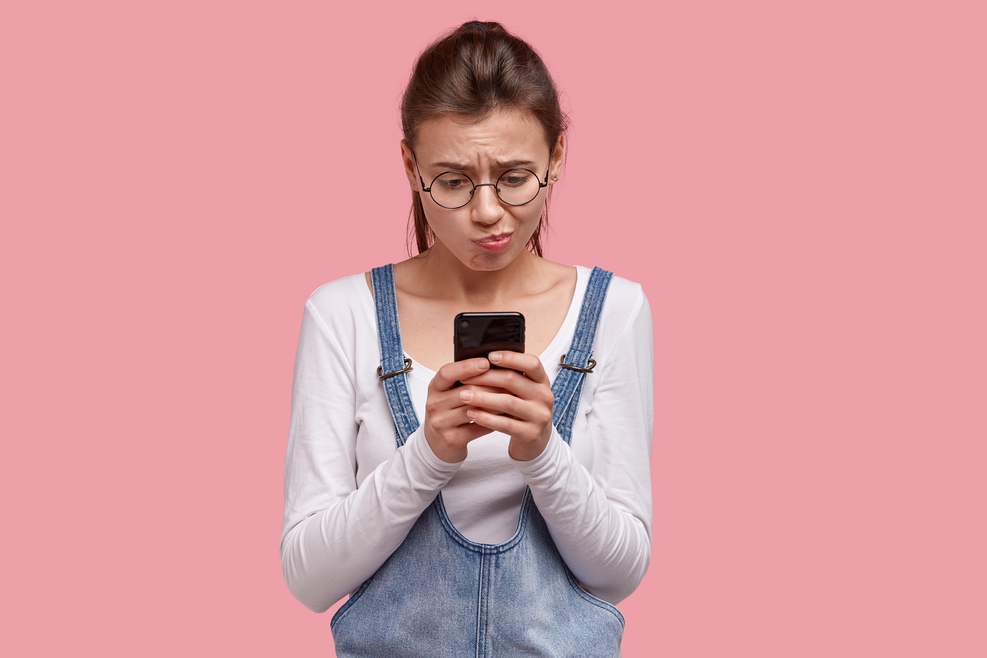 unhappy lady holds smart phone, has dissatisfied expression as battery died, cant watch video, wears round spectacles, denim sarafan, poses over pink background, has negative opinion about something
