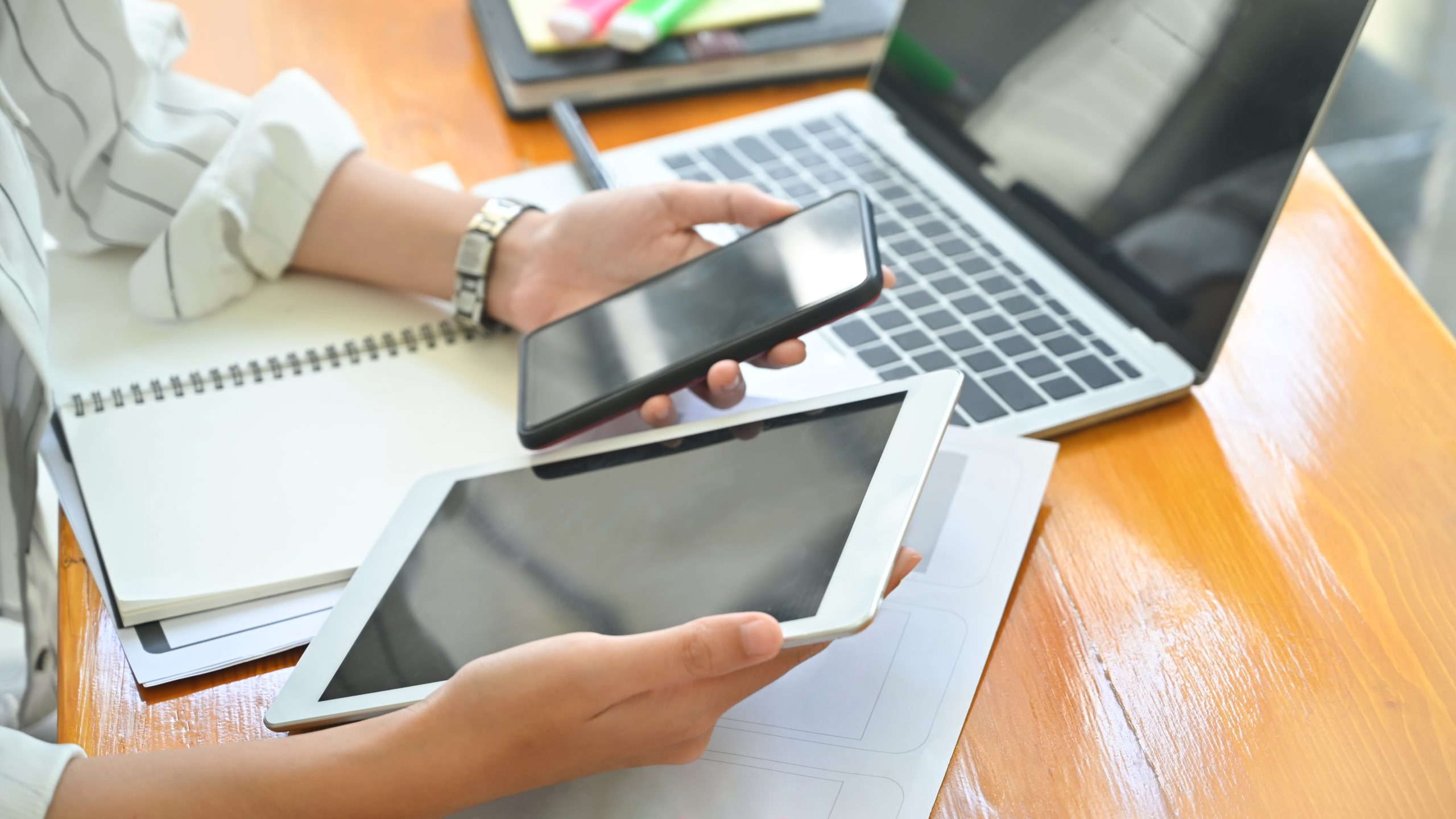Profissional segurando e comparando celular e tablet sobre a mesa de trabalho ao lado do notebook