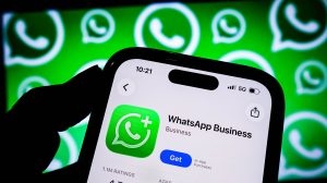 Mão segurando celular com a tela do aplicativo WhatsApp Business, com fundo de logotipos do WhatsApp